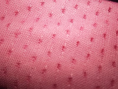 A close-up, macro view of a pink woven fabric is depicted, located in Orifite, Nigeria. The textile surface exhibits a fine, regular weave with numerous small, irregularly shaped, darker red raised dots or flecks distributed uniformly across its area. The primary color is a mid-tone pink, with the dots providing a darker contrasting hue and tactile texture. The image demonstrates a shallow depth of field, rendering some regions of the fabric slightly out of focus. No individuals, distinct objects beyond the fabric, or specific environmental markers are discernible within the frame.