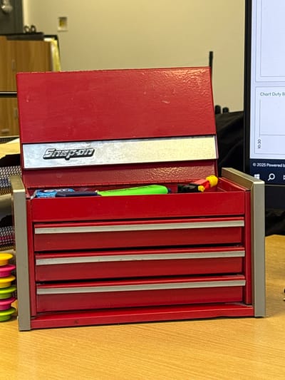 A red Snap-on branded miniature toolbox, with its lid open, sits on a light-colored wooden desk surface in an office environment in Nibley, United Kingdom. The toolbox features a silver band with the "Snap-on" logo below the lid and four red drawers with silver-colored pull strips. Inside the open upper compartment, a blue object, a bright green highlighter, and a small yellow and red toy figure are visible. To the left of the toolbox, a black wire mesh organizer contains stacked circular objects in yellow, pink, and green. In the background to the left, a light-colored wooden cabinet is partially observed. To the right, a computer monitor displays a chart interface with text "Chart Duty B..." and a "© 2025 Powered by..." copyright notice. A dark object is partially visible behind the monitor.