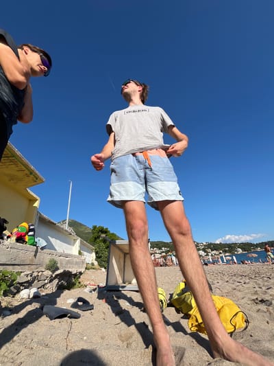 Two young men are on a sandy beach in Zankovići, Montenegro. The man on the left, partially visible and wearing sunglasses, looks down. The man on the right, in focus and also wearing sunglasses, looks up at the sky. He is wearing a grey t-shirt with "adidas" printed on it and light blue and light grey swim shorts. He is adjusting his shorts. A bright yellow bag lies on the sand next to his feet. The background shows a clear blue sky, a building with a yellow awning, a lamp post, vegetation, and the coastline with buildings and people.