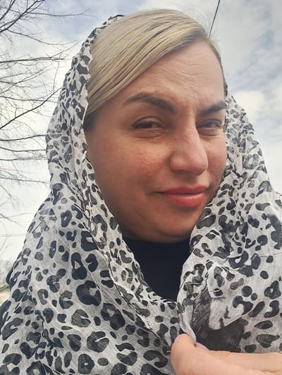 A close-up, low-angle portrait depicts a fair-skinned woman with blonde hair, observed outdoors in Avdot’ino, Russia. Her blonde hair is pulled back, partially covered by a light-colored sheer scarf featuring a black leopard print pattern. She wears a dark garment underneath the scarf. The woman is looking directly towards the camera with a slight smirk; her eyes are partially squinted. Her right hand, featuring light pink nail polish on the thumb, is visible at the bottom right, gently holding a fold of the scarf. The background consists of out-of-focus bare tree branches against a cloudy or overcast sky.