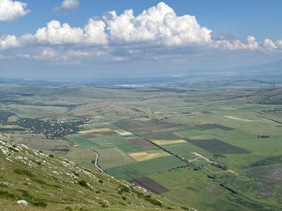 The image displays a panoramic aerial view of a rural landscape from a high vantage point, likely a mountainside. In the immediate foreground, a steep grassy slope covered with numerous white rocks descends towards the midground.
The midground features a diverse mosaic of agricultural fields, forming a patchwork of greens, yellows, and browns, indicative of different crops and soil conditions. A winding, paved road is visible, leading towards a village situated in a valley on the left side of the frame. The village consists of numerous buildings with reddish roofs, densely interspersed with trees. A distinct, linear feature, possibly a path or field boundary, runs horizontally through the center of the cultivated fields. Scattered clusters of trees are present throughout the midground.
Beyond the cultivated fields, the terrain transitions into rolling, vegetated hills that extend into the background. A large, light-colored patch of land, possibly a quarry or cleared area, is discernible in the mid-distance. Further in the background, a distant urban area with a skyline of buildings is faintly visible on the horizon, partially obscured by atmospheric haze.
The sky above is a clear blue, adorned with numerous white, puffy cumulus clouds, with a prominent cluster dominating the upper-central portion of the frame. The overall lighting suggests bright daylight. No people or animals are present in the scene. The image captures a broad rural vista in the vicinity of Dragoman, Bulgaria, characterized by agricultural activity and hilly topography.