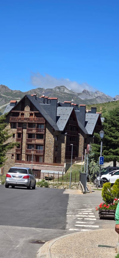 The image depicts a multi-story stone building with dark slate roofs, nestled against a backdrop of lush green mountains under a clear blue sky. The building appears to be a hotel or residential complex, featuring numerous balconies and large windows. In the foreground, a silver car is parked on a paved road. A pedestrian crossing is marked on the road with white stripes. A blue sign with a wheelchair symbol indicates accessibility. The scene suggests a daytime setting with fair weather. The overall ambiance is serene, possibly indicating a tourist destination in a mountainous region. The text "CU 615 SA" is visible on the license plate of the silver car. The location is identified as Sallent de Gállego, Spain.