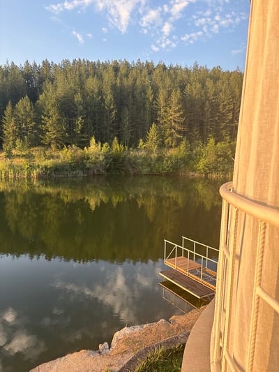 The image depicts a tranquil lake scene in Skalsko, Bulgaria, with a dense pine forest lining the opposite shore. The water perfectly reflects the trees and the blue sky dotted with white clouds. In the foreground, a small wooden dock with white railings extends into the lake. To the right, a section of a light-colored, translucent curtain and a white metal railing are visible, suggesting a structure adjacent to the water's edge. The lighting indicates it is either early morning or late afternoon, with the sun casting a warm glow on the scene. There are no people present in the image.