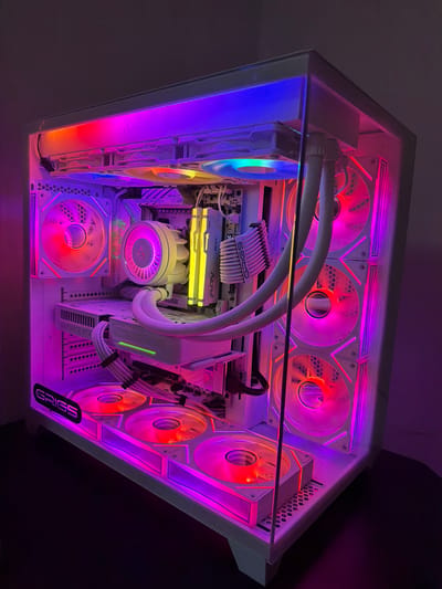 The image shows the interior of a custom-built computer tower. The components are lit with RGB lighting, cycling through various colors, including blue, purple, pink, orange, and yellow. Visible components include multiple cooling fans (top, side, bottom), a water cooling system with tubes and radiator, RAM sticks labeled "FURY BEAST", a GPU, and custom white cables. The case itself is white and has the word "GRIGS" printed on the bottom right corner. The location in the city of Dragor, Bulgaria is not discernible from the provided image.