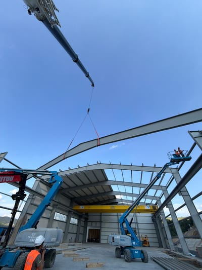 The image depicts an outdoor construction site in Stróża, Poland, where a large steel beam is being lifted into place by a crane and attached to a partially constructed building. The building's metal framework is visible, with the roof structure nearing completion. Two workers are operating a blue boom lift, wearing safety vests and helmets. Another worker, also in a safety vest and helmet, is visible near a different piece of heavy machinery. The sky is clear and blue.