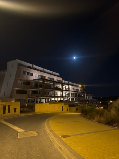 This is a night-time exterior shot of a modern building in Finestrat, Spain, possibly an apartment complex or hotel. The building features a white facade on the left and beige, wood-like paneling on the right. It has multiple levels with balconies and dark windows. The scene is illuminated by artificial lights from street lamps and within the building, as well as the glow of a full moon in the dark sky. A paved road curves in the foreground, with yellow brick paving along the right side. There are some low walls made of yellow stone blocks. The overall environment appears to be a developing or residential area with some sparse vegetation.