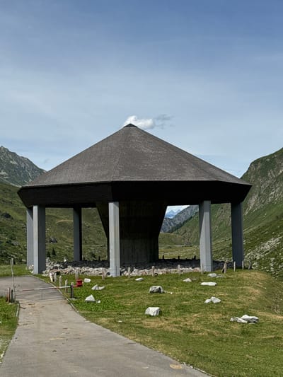 The image shows a modern, angular building with a dark, textured roof supported by large, grey pillars. The building is situated on a grassy mountainside, with a paved pathway running alongside it. In the background, a dramatic mountain landscape unfolds under a bright, clear blue sky with some wispy clouds. The green slopes are dotted with rocks and small patches of vegetation. The sunlight suggests it is daytime. The overall scene evokes a sense of tranquility and awe at the natural beauty of the mountainous region, possibly a viewpoint or a rest stop for hikers. There are no people visible. The building's design is stark and geometric, contrasting with the organic forms of the surrounding environment.