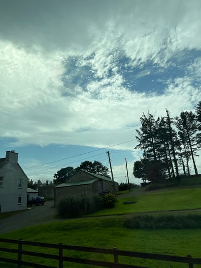 This is a landscape photograph taken in Massinass, Ireland. The image is oriented vertically, with the sky occupying the left half of the frame and the ground the right. Wispy white clouds are scattered across a pale blue sky, suggesting a partly cloudy day. Tall, dark green pine trees line the horizon on the left side, with their branches silhouetted against the sky.

In the mid-ground, there are several buildings. A long, low structure with a corrugated roof is visible, and next to it, a smaller building with a stone facade. A dark-colored car is parked on a gravel driveway leading towards these buildings. Further in the foreground, a light-colored two-story house with white-framed windows is partially visible. A dark wooden fence with horizontal rails runs along the right edge of the image, separating the property from a lush green field. The field appears to be a well-maintained lawn, with some areas of shadow indicating uneven terrain or tree cover. Overhead, power lines stretch across the frame, supported by utility poles. The overall impression is one of a rural or semi-rural setting, possibly a farm or a residential area with agricultural land nearby. The time of day appears to be daytime, with diffused light suggesting it might be overcast or late afternoon. No text is visible in the image.