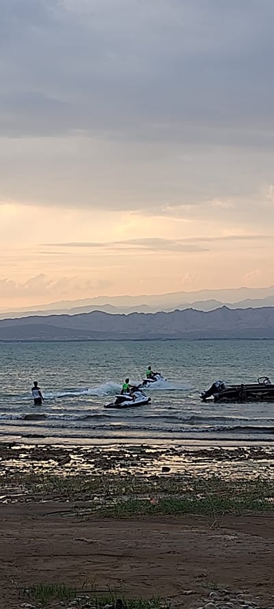 The image depicts a serene body of water with a distant mountainous horizon under a cloudy sky. In the foreground, a sandy shore is visible, with some sparse vegetation and debris. The water's surface is rippled, reflecting the subdued light. Several jet skis are in motion on the water. Two jet skis are actively being ridden by individuals wearing green tops. One jet ski is positioned slightly ahead of the other, creating a wake. A third jet ski, riderless, is moving further out. To the left of the jet skis, a person stands in the shallow water, facing away from the camera. To the right, a portion of a boat with an outboard motor is partially submerged in the water. The scene suggests a recreational activity taking place on a lake or sea near Chigdalik, Tajikistan, with the mountains providing a backdrop to the water activities.