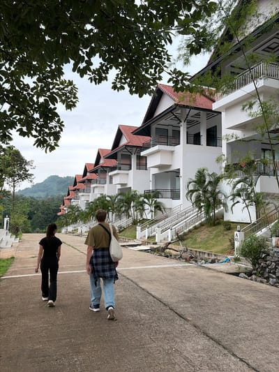 In Ban Kao Dua, Thailand, two individuals are walking away from the camera on a concrete pathway. The person on the left wears a black t-shirt and black pants, while the person on the right is dressed in a khaki t-shirt, blue jeans, and a plaid shirt tied around their waist. They are walking towards a row of modern white buildings with red tiled roofs, characteristic of resort-style architecture. The buildings are situated on a gentle slope, with multiple levels and balconies. Lush green trees and foliage surround the buildings, and a mountainous landscape is visible in the background under a cloudy sky. Some of the buildings have visible staircases and railings, and landscaping includes palm trees and grassy areas.