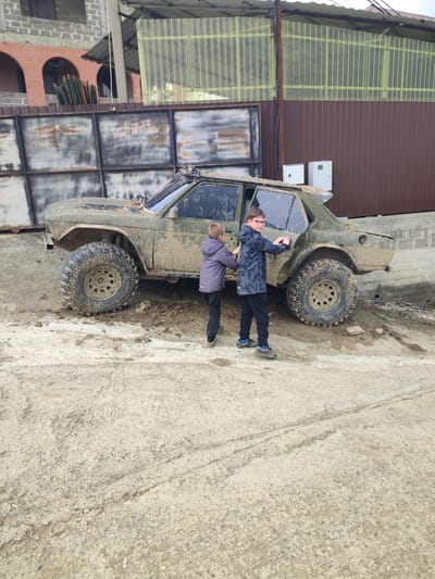 The image displays two young boys attempting to move a heavily mud-covered, olive green off-road vehicle in an outdoor setting within Bestuzhevskoye, Russia. The precise location within the city is not discernible from the visual information provided, but the environment suggests a semi-rural area, a private yard, or an unpaved access road adjacent to various structures.

The vehicle is a modified four-door sedan, extensively coated in dark brown mud across its body, windows, and large, knobby off-road tires. It appears to be partially stuck, with its rear driver's side wheel and front left wheel embedded in a significant accumulation of mud.

Two boys are positioned next to the vehicle's rear passenger side. The boy on the left, dressed in a grey and purple hooded jacket and dark pants, is pushing against the rear passenger door with both hands. The boy on the right, wearing glasses, a dark camouflage-patterned hooded jacket, and dark pants with blue and black athletic shoes, is similarly pushing against the same door with both hands, looking toward the left side of the frame.

The ground directly in the foreground consists of a light grey, coarse concrete or hardened dirt surface exhibiting tire tracks, transitioning into the muddy patch where the vehicle is situated. In the background, a tall, rusty, square-paneled metal fence serves as a boundary. Behind this fence, on the left, a partially constructed building made of grey concrete blocks with red brick arched openings is visible, alongside stacked green objects. To the right of the fence, a brown corrugated metal wall extends upwards, topped by a translucent corrugated roof structure. Two small, rectangular white electrical boxes are affixed to the brown wall. The sky is overcast.