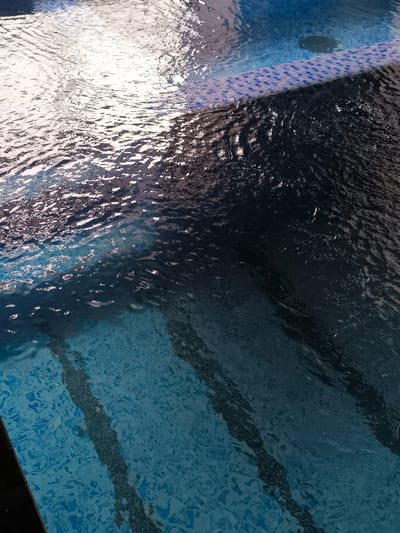 The image depicts a close-up, high-angle view of a swimming pool located in Charvak, Uzbekistan. The pool water is clear, exhibiting ripples on its surface that distort reflections of overhead lighting. The pool bottom and visible side are tiled with light blue mosaic squares, forming a textured pattern. A distinct line of darker blue mosaic tiles marks what appears to be an edge or step within the pool. A circular drain or skimmer cover is visible in the darker tiled section near the water's surface. Shadows are cast beneath the water, indicating the presence of underwater features or variations in depth. No individuals, actions, or specific objects beyond the pool infrastructure are visible.