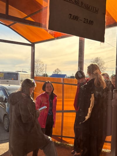 A daytime shot shows an outdoor location in Gory, Russia. Two females are standing outside a building, reflected in the glass. A sign on the building states "HOURS OF OPERATION" in Russian, with the hours listed as "7.00-23.00." A vehicle with a school zone symbol on the back is parked nearby. The weather appears sunny.