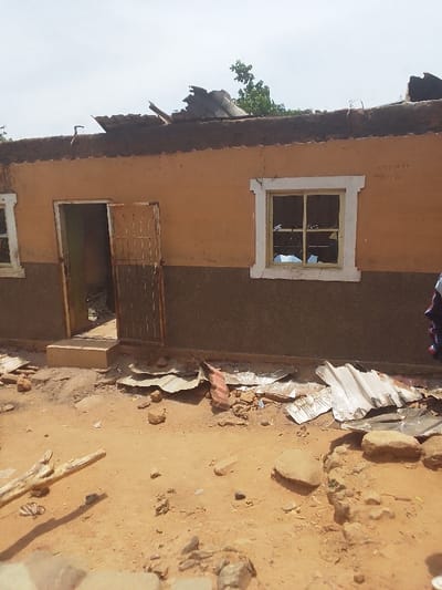 The image shows a damaged building located at coordinates 9.804, 8.646 in Kwal, Nigeria. The building is made of brick and is painted a light orange color. The roof is partially destroyed with pieces of corrugated metal lying on the ground around the building. There is a window on the right side of the building that is also broken and a closed door on the left side of the building. The door is made of metal and has a barred opening. There are also a few rocks and pieces of wood scattered around the building. The ground is dirt and there is a small amount of vegetation in the background. There are no people present.