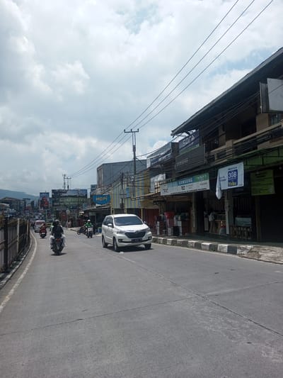 The image depicts a street scene, likely in Southeast Asia, characterized by urban activity and commercial presence. A white car is driving on the right side of the road, with two motorcycles traveling in the opposite direction. Buildings line the street, adorned with various commercial signs advertising brands like Samsung, OPPO, and Xiaomi. A sign mentioning "3GB 11ribu" suggests a promotion for mobile data.
The sky is overcast, and utility poles with electrical wires are visible overhead. A fence runs along the left side of the road. The road itself shows signs of wear and tear, with patched areas and visible seams. The environment appears to be a mix of residential and commercial properties. A banner with the words "JAYA Ad" and several phone numbers can be seen in the background.