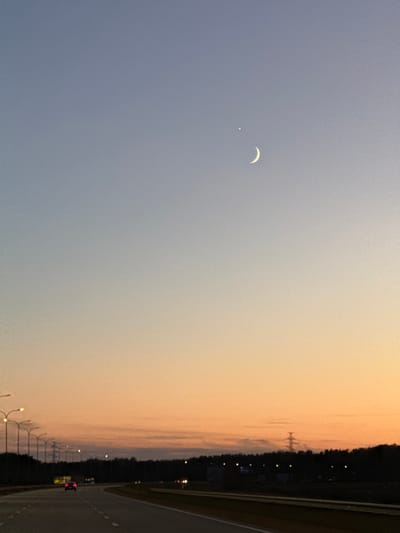 The image depicts a twilight scene on a highway, likely in or near Piņķi, Latvia. The sky transitions from a pale blue at the top to a warm orange and yellow gradient near the horizon. A crescent moon and a bright star are visible in the upper portion of the sky. The foreground shows a dark asphalt highway with white lane markings. On the left side of the highway, a row of streetlights with glowing bulbs lines the road. In the distance, a dense line of dark trees forms the horizon. A power line tower can be seen silhouetted against the sky above the trees. Several cars are visible on the highway, with headlights and taillights illuminated. One red car in the middle distance is particularly noticeable. The overall impression is one of a serene, late evening drive.