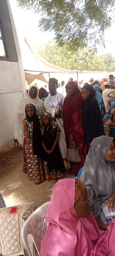 This image captures a daytime outdoor gathering in Dorayi, Nigeria. A prominent group of seven individuals, comprising three adult women, two young girls, and one young adult male, stands in the midground, posing for a photograph. The young male, positioned centrally, wears a light-colored traditional outfit, a patterned cap, and a white sash with blue Arabic script, holding a blue ornate object. The two girls wear black headscarves and similar patterned dresses featuring circular designs. An adult woman to the left wears a white hijab and garment. To the male's right, an adult woman in a dark red hijab and dress carries a beige structured handbag. Another adult woman to her right wears a dark blue hijab and dress. The group stands near the side of a white vehicle, possibly a bus or van. The ground consists of dry earth. In the background, numerous other individuals, predominantly women in traditional attire and head coverings, are visible seated and standing under makeshift tent-like structures, with green foliage from trees and a bright sky overhead. In the immediate foreground, a woman wearing a pink hijab is partially visible, seated on a white plastic chair.