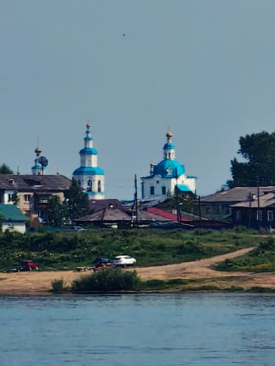 A distant, slightly out-of-focus view of the city of Yeniseysk, Russia, showcases a waterfront scene. The foreground features a body of water, likely the Yenisei River, with ripples suggesting a gentle current. Along the riverbank, a sandy shore is visible, with a path winding through green vegetation and some bushes.
Further inland, a cluster of buildings forms the cityscape. Dominating the view are two distinctive churches with white walls and vibrant blue domes, topped with golden crosses. One church is taller and more prominent, featuring a bell tower. Adjacent to the churches are residential buildings with darker roofs and visible windows. Some of these appear to be older, wooden structures, while others are more modern.
On the sandy bank, two vehicles are parked. A red car is on the left, seemingly attached to a trailer. To its right, a white car is also parked, also with a trailer. The overall impression is one of a peaceful, somewhat rural urban setting. The sky is a pale blue, with a single small dark speck visible high up, possibly a bird or an insect.
