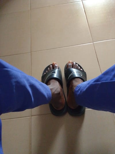 The image shows a close-up view of a person's feet wearing black Nike slide sandals, with their legs covered by blue trousers. The feet are positioned on a tiled floor, which appears to be light brown or beige. The lighting suggests it is daytime, and there are no visible indications of weather conditions. The scene is simple and focuses on the footwear and clothing. The background consists of the tiled floor, which shows grout lines dividing the tiles. The image is taken from a slightly elevated perspective, looking down at the feet. There is no discernible text in the image. The overall impression is a casual and everyday moment, possibly taken indoors or in a sheltered outdoor area. The location context provided is Abeere, Nigeria.