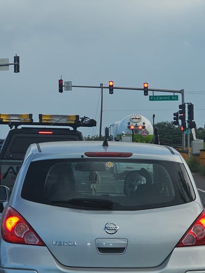 The image depicts a traffic scene at an intersection. In the foreground, the rear of a silver Nissan Versa is visible, with its taillight illuminated. A propane tanker truck is positioned behind the car, its large white tank prominently displayed with green accents and markings. The truck has the number "1075" and "500" on its side, along with the word "PROPANE." The scene appears to be set during the day, under an overcast sky, as indicated by the flat, diffused lighting and the greyish sky.
The intersection features multiple traffic lights, some of which are red, suggesting a stop or waiting period. A street sign indicates "FLEMING Rd." A "LEFT ON ARROW ONLY" sign is also visible. The overall setting appears to be a suburban or semi-industrial area, with trees visible in the background. The mood of the image is neutral, reflecting a typical traffic situation. There are no visible people, but a reflection in the Nissan Versa's rear window shows what might be a person's face or a reflection of an object.