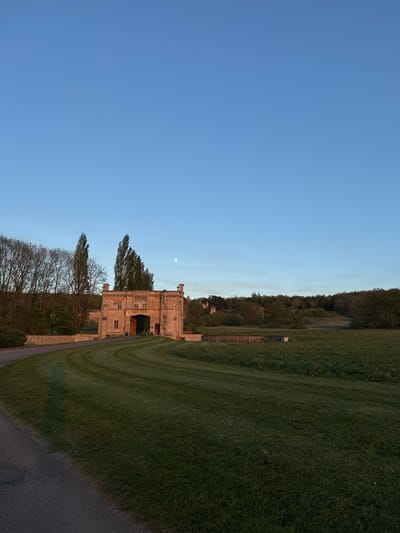 The image displays a picturesque landscape centered around a grand, historic building. The edifice, constructed from light-toned stone, features multiple stories, numerous windows, and intricate architectural details on its roofline, suggesting a manor house or stately home. It is partially framed by a line of trees, some with bare branches indicating a seasonal transition, while others are verdant.
A vast, well-manicured green lawn occupies the foreground and mid-ground, showing distinct mowing patterns. A paved path or road runs along the lower right edge and curves into the left foreground. In the background, beyond the building and grounds, a dense, dark green wooded hillside rises, providing a natural backdrop to the scene.
The sky above is a clear, bright blue, devoid of clouds. A prominent, pale moon is visible high in the sky, positioned to the left of the building's peak. The presence of the moon alongside a bright sky suggests the photograph was taken during either early morning or late afternoon. The lighting is soft and natural, contributing to a tranquil atmosphere. There are no people visible in the image, nor is any text discernible. The scene evokes a sense of historical elegance and serene natural beauty within a rural or parkland setting.