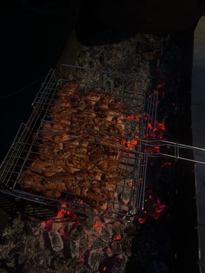 This media file depicts a grilling basket filled with numerous pieces of seasoned meat, likely chicken or pork, being cooked over hot coals and embers in an outdoor setting. The meat is browned and appears to be well-marinated, with some pieces exhibiting a glossy sheen from the cooking process. The grilling basket itself is made of wire mesh and has a handle. The background shows a fire pit filled with glowing red coals and ash, indicating the heat source. The overall scene suggests a casual outdoor meal or gathering. The lighting is dim, with the glow of the coals providing illumination, suggesting it is either evening or nighttime. The location context provided is Solontsy, Russia, which implies a potentially rural or suburban environment. There is no visible text in the image. The activity taking place is the grilling of meat, a common practice for outdoor cooking and social events. The image evokes a sense of warmth, anticipation of food, and potentially relaxation associated with a barbecue.