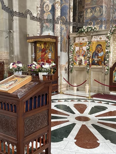 This appears to be the interior of the Manasija Monastery located in Despotovac Municipality, Serbia. The image shows various religious icons adorned with flowers, a wooden pulpit with intricate carvings, and a decorative marble floor. The walls and ceilings exhibit Byzantine-style frescoes. There are no people visible in the frame.