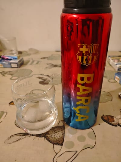 A close-up shot of a cluttered table. A clear glass with ice sits to the left, partially filled with water. To the right is a metallic water bottle, red at the top and fading to blue at the bottom, adorned with the FC Barcelona logo and the word "BARCA" in yellow. Cigarette packs are scattered around the glass and bottle. The table is covered with a patterned tablecloth featuring drawings of cups. The location in Swindon, United Kingdom, cannot be determined from the image alone.