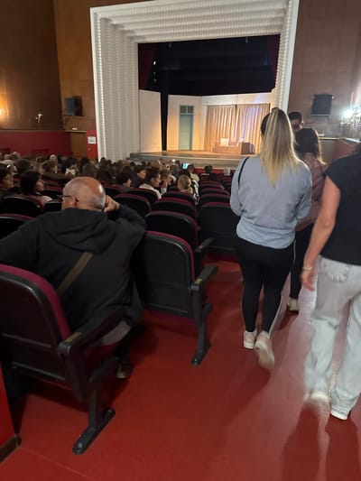 This image depicts an indoor scene within an auditorium or theater, located in Slivnitsa, Bulgaria. The perspective is from an aisle, looking towards the stage area on the left and across rows of seating on the right.
The primary subjects include several individuals. In the foreground/mid-ground, a woman with long blonde hair, wearing a light blue top and black leggings, is walking away from the camera. Behind her, another person in a black top and light-colored pants is also in motion. To their right, a bald man wearing glasses and a dark jacket with a brown stripe is seated in an aisle seat, appearing to be resting or looking down. Further into the background, numerous other individuals are seated in rows of theater seats, indicating an audience.
The setting is characterized by a red carpeted floor and rows of dark red and black theater seats. On the left side of the image, parts of the stage or performance area are visible, featuring white ornate architectural moldings and dark curtains. Artificial lighting fixtures are visible overhead. A light blue framed structure, possibly a window or decorative panel, is also present on the left wall.
The scene suggests an event is either about to begin, is in progress (with some attendees moving), or has recently concluded. No specific text is discernible within the image.