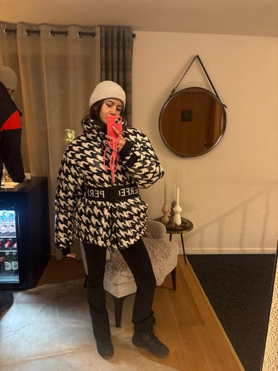 A person wearing a black and white patterned jacket, black pants, boots, and a beanie takes a selfie with a pink phone. Behind the person, there's a mirror, a chair with a fuzzy cushion, and a candle display on a small table. Curtains are visible on the left. A mini fridge displaying various bottles and cans is in the foreground. There is another person in the reflection.
