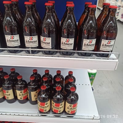 The image shows a close-up view of several bottles of Ichnusa beer arranged on supermarket shelves. The bottles are dark brown glass with red caps and feature distinctive yellow and red labels. The labels prominently display the "Ichnusa" brand name, along with text indicating "AMBRA LIMIPIDA" and "NON FILTRATA" on some bottles. The percentage "5% vol." is also visible on the labels. Some bottles show the vintage "1912". The bottles are positioned on two shelves, with a metal railing in the foreground. In the background, out-of-focus items include more bottles and what appears to be a roll of paper product. The lighting is bright, suggesting an indoor retail environment. The timestamp "26.06.2026 09:52" is superimposed on the lower right corner of the image, indicating the date and time the photograph was taken. There is no visible indication of the specific location within Terlizzi, Italy, other than the presence of a retail store.