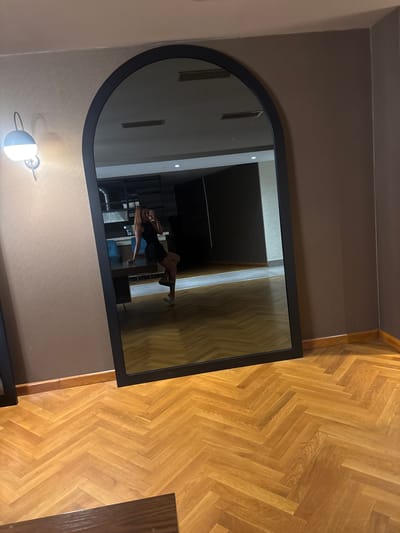 A full-length mirror with a black frame is positioned against a taupe-colored wall. The mirror reflects a woman in a black dress and white shoes, posing by sitting on a desk. She is holding a phone and taking a selfie. The room has a herringbone-patterned wooden floor and a single wall lamp on the left side of the frame. The background visible in the mirror shows a modern interior with furniture and other reflective surfaces. The image appears to be taken in a residential or commercial space in the Kâğıthane district of Istanbul, Turkey.