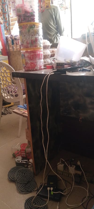 This image depicts the interior of a small retail establishment in Danbare, Nigeria, likely a convenience store or bakery.

In the foreground, a dark-colored counter or desk runs horizontally, with white charging cables dangling over its edge. To the left of the counter is a white plastic chair featuring a pattern of circular cutouts. On the light-colored tiled floor, several coiled grey objects, resembling mosquito coils, are arranged. Adjacent to these coils are two black power strips connected by various white and black cables, some plugged into wall adapters. A silver padlock and a clear plastic bottle with red contents are also visible on the floor near the coiled objects.

On the counter, a tall stack of clear plastic containers, each filled with colorful items such as sweets or snacks, is prominently displayed. To the right, a translucent white plastic container rests on the counter, partially obscuring other items.

In the background, a person, seen from behind, wears a light green long-sleeved shirt and stands facing a brightly lit window or doorway, which appears to be the primary light source. To the left of this person, shelves stocked with various packaged goods, including bread and possibly biscuits or other snacks, are visible. Below these shelves and behind the stack of clear containers, multiple clear plastic clamshell containers filled with baked goods, such as buns or cakes, are stacked.