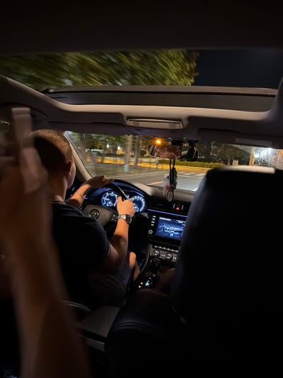 The image shows the interior of a car at night, viewed from the back seat. A person is driving, with their hands on the steering wheel. The dashboard and a large infotainment screen are visible. Outside the car, trees and streetlights can be seen. The car has a sunroof open, and the sky is dark. The reflection in the rearview mirror shows the driver's face. There is also a car air freshener hanging from the mirror. The scene appears to be on a city street. Based on the context provided, this is likely in Brno, Czechia.