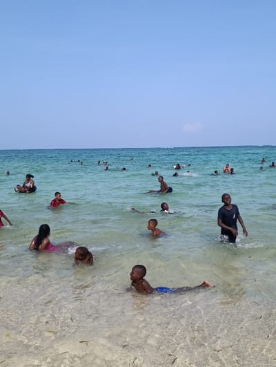 A sunny beach scene shows many people, mostly children, enjoying the shallow, clear ocean water. Some are swimming, wading, or floating. The water is calm and light turquoise, fading to a deeper blue further out. The sandy beach is visible in the foreground. The sky is clear blue. The overall impression is of a joyful and relaxed atmosphere.