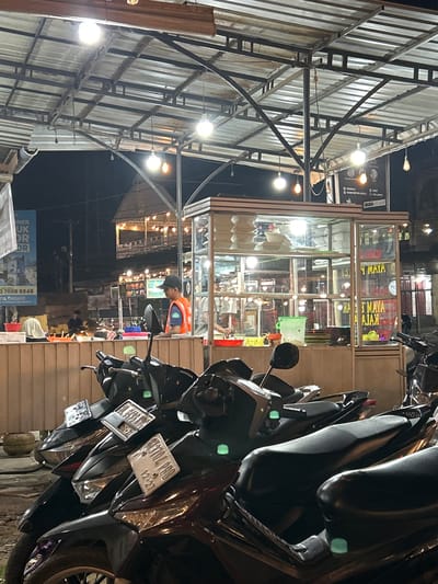 A nighttime outdoor food stall scene is depicted in Tambonbaroh, Indonesia. Several motorcycles are parked in the foreground, facing left. Behind them, a wooden counter with a glass display case is set up. Inside the case, food items are visible, and a person wearing an orange vest and a dark cap stands behind the counter, appearing to be working. The stall is illuminated by overhead lights, and the background shows a street scene with more lights and structures.