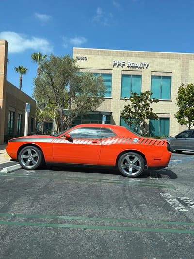 The image depicts an orange Dodge Challenger parked in a lot in Irvine, California. The car is positioned in a parking space marked with green lines. Behind the car is a building with "PPF REALTY" signage and an address number "16450". Palm trees and other foliage are visible around the building and parking lot. The sky is blue with some scattered clouds. A portion of another vehicle, a silver sedan, is visible to the right of the Dodge Challenger. The ground of the parking lot is asphalt with white painted markings, some of which indicate "RESERVED". A blue handicapped parking sign is visible to the left of the Dodge Challenger.