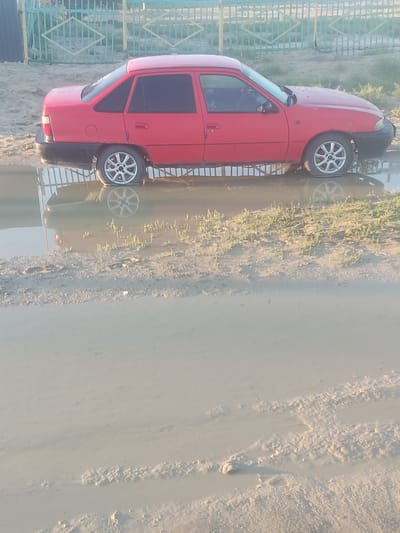 A red sedan is partially submerged in a large puddle of muddy water on an unpaved road. The car appears to be stuck, with its wheels deep in the water. The setting is a rural or semi-rural area, suggested by the unpaved road and the fence in the background. The fence is a light green metal fence with diamond-shaped openings. The ground is dirt and mud, and some sparse green vegetation is growing in patches. The sky is overcast, casting a soft light on the scene. The image was taken in the city of Aral, Kazakhstan.