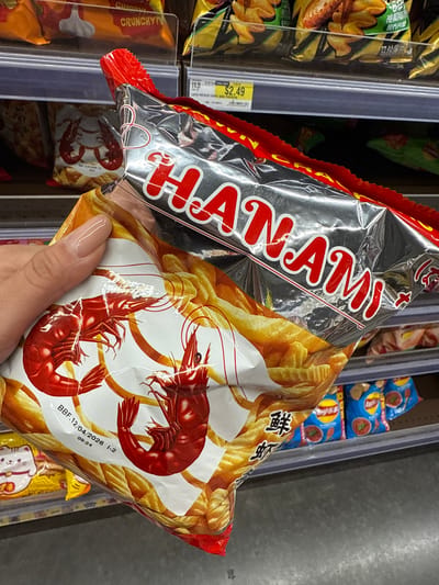 A hand holds a bag of Hanami brand prawn crackers in a store, likely a supermarket, with shelves of other snack foods visible in the background. The packaging is predominantly red, silver, and white, featuring images of prawns and what appears to be crunchy chips. The Best Before date on the bag reads "BBF.12/04/2026". A price tag showing "$2.49" can be seen on a nearby shelf. The location is likely Orlando, United States, based on the prompt.
