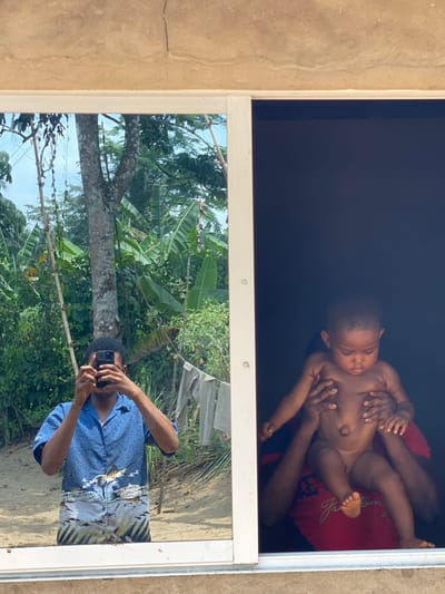 In Ikot Ete Ekpo, Nigeria, the image presents a bifurcated scene observed through a white-framed window. The upper section reveals a light brown, rough-textured concrete wall with minor cracks.
The left pane of the window functions as a reflective surface, showing the outdoor environment and the photographer. A dark-skinned adult, presumably male, is reflected from the chest up, holding a dark smartphone directed towards the scene, thus capturing their own image. He wears a short-sleeved blue collared shirt with a light-colored graphic depicting ocean waves and a fish. Behind the reflected individual, a lush, tropical outdoor setting is visible, characterized by prominent green foliage, large-leafed plants resembling banana trees, tree trunks, and a sandy or dusty ground. Light-colored laundry is seen hanging on a line or bush in the background.
The right pane of the window offers a direct view into a dark, interior space. A dark-skinned infant, appearing unclothed, is held upright by hands belonging to an individual partially visible below. This individual wears a red short-sleeved t-shirt with a white graphic or text element. The infant looks downwards, and the interior background is dimly lit.