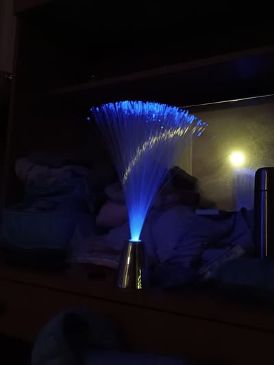 A fiber optic lamp with blue light is on a shelf or table. The room is dark, with some items stored on the shelf behind the lamp, including a metallic thermos. A light source is visible on the wall behind the lamp. The location is Cherkovitsa, Bulgaria.