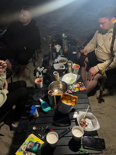 The image depicts an outdoor gathering on a sandy surface, likely a beach, at night. Three individuals are present, two men and one person whose gender is not clearly discernible due to their clothing and pose. The men are seated at a black picnic table laden with food and drinks. One man, wearing a black hoodie, is seated on the left, looking away from the camera. Another man, in a tan hoodie, is seated on the right, holding a smartphone. A third individual, wearing a light-colored hoodie and black pants, is seated on the left, partially visible.
On the table, there are several containers of food, including a bowl of chips, a bowl with remnants of food, and several smaller containers holding what appears to be sauce or dips. Various beverages are also present, including cans of beer and bottled drinks. Other objects on the table include a pair of sunglasses, a lighter, a bottle opener, keys, and a smartphone. A portable stove with a burning element is also visible on the table. The scene is illuminated by artificial light sources, creating shadows and highlighting the sandy environment. The overall impression is that of a casual outdoor meal or gathering. The location is identified as Povarovka, Russia.