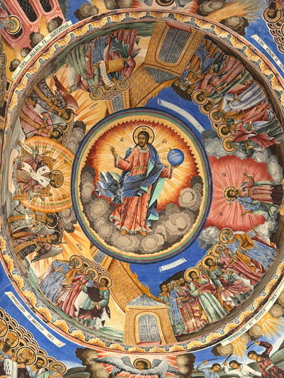 This is a highly detailed, circular fresco painting. The scene is complex, featuring numerous figures arranged in concentric circles within a dome-like structure.
In the central part of the fresco, a figure lies horizontally, draped in blue and red robes, holding a blue orb above. The figure is surrounded by grey clouds and radiating yellow light.
Around this central scene, a second ring depicts various figures and events. One section shows a seated, bearded figure on a throne, flanked by angel-like beings. The rest of the ring contains groups of people.
The outermost ring shows individuals, primarily depicted from the chest up. Each has a halo and appears in various poses. This outermost ring is bordered by a band of text in a Slavic language. The background colors shift from blue to reddish-orange to yellow.
The style is reminiscent of Byzantine art with stylized figures, flat perspective, and rich, symbolic colors.
The location is likely within the Rila Monastery (Rilski manastir) in Bulgaria, a place known for such religious frescoes.