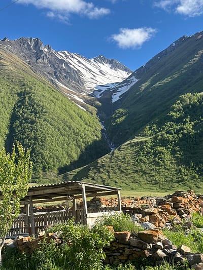 This photograph depicts a mountainous landscape in Ketrisi, Georgia, under a bright blue sky with scattered white clouds. The foreground features a rustic wooden structure, possibly a shed or shelter, with a corrugated metal roof. Adjacent to it, a fence made of vertical wooden planks encloses an area. A pile of reddish-brown stones is visible to the right of the structure, and further to the right, more stones are stacked in a rough wall. Green vegetation, including tall grass and leafy plants, grows around the stones and in front of the fence. To the left of the wooden structure, a leafy green tree stands.

The midground consists of lush green hillsides, densely covered with trees. These hills rise steeply, leading to the majestic mountains in the background. The mountains are characterized by rugged, rocky peaks, with patches of snow clinging to their upper slopes, particularly in the central and left sections. A narrow, winding ravine with a visible watercourse, possibly a stream or small waterfall, descends from the snowy peaks through the green slopes. The overall environment suggests a rural, mountainous setting. No people are present in the image.