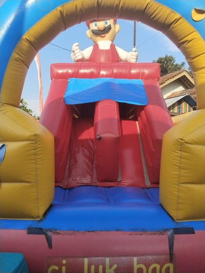 A colorful inflatable bounce house stands outdoors in Sambirejo, Indonesia. It features a Mario character figure at the top of the slide. The bounce house is constructed with blue, yellow, and red vinyl. A blue canopy covers the slide entrance. The surrounding area appears to be a residential neighborhood with houses and trees visible in the background.