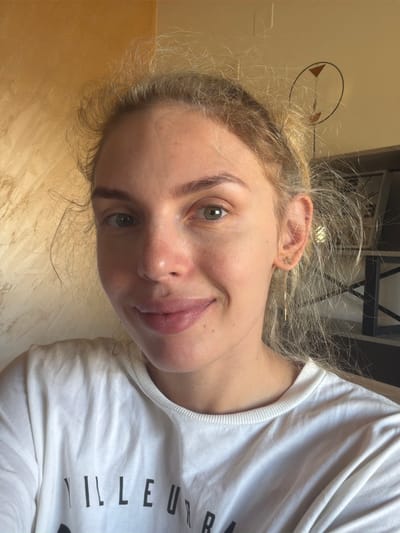 This is a close-up, frontal selfie of a young Caucasian woman with blonde hair, taken indoors. She is wearing a white t-shirt with black text on the front. The woman has light skin with freckles, green eyes, and is smiling. She is wearing a gold earring in her right ear. The background appears to be a wall with a textured finish. The lighting suggests natural light is coming from the right side of the frame. The scene is located in Mojanovići, Montenegro.