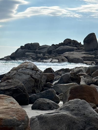 The image shows a rocky coastline near Hout Bay, South Africa. Large, gray rocks and boulders dominate the foreground and extend along the coastline. The ocean is visible between the rocks, with waves crashing against them. The sky is pale blue with some white clouds. The overall scene depicts a rugged and natural environment. There are no people visible.