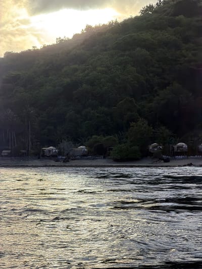 A body of water, shimmering with reflected sunlight, fills the right half of the image. On the left, a densely forested hill slopes downward to meet a narrow strip of beach. Small structures, possibly huts or buildings, are spaced along the beach at the edge of the forest. The sun is obscured by clouds, casting a soft glow on the scene. The perspective is tilted, giving a somewhat disorienting view.