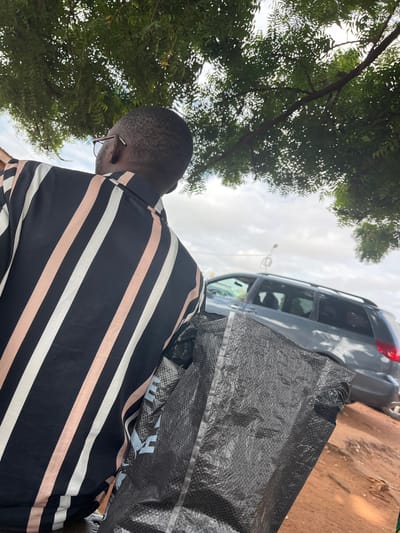 The image depicts an outdoor scene in Idah, Nigeria, viewed from slightly behind and below a person. A person with short, dark hair, wearing brown-framed eyeglasses, is seen from the back, facing towards the right side of the frame. They are wearing a black shirt with distinct vertical stripes in white and light peach/pink colors. The person is carrying a large, black, textured woven plastic bag, possibly a polypropylene sack, which occupies a significant portion of the lower right foreground. Light blue markings are faintly visible on the bag's surface.
In the mid-ground, a dark gray minivan or SUV with roof rails is partially visible, positioned on a reddish-brown dirt or soil surface that suggests an unpaved area or roadside. The vehicle's rear section, including a portion of its red taillight, is visible on the right.
The upper portion of the image is dominated by dense, green foliage from a large tree, with patches of bright, overcast sky visible through the leaves. The sky is predominantly cloudy with diffused light. A slender, light-colored pole or structure is faintly discernible in the distance beyond the vehicle. The overall lighting suggests natural daylight under a cloudy condition.