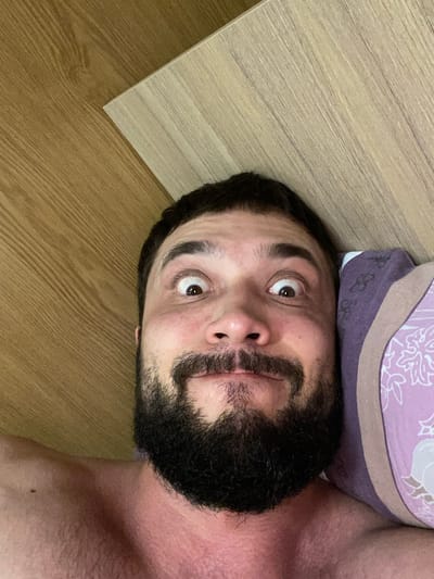 The image is a close-up selfie of a man with a beard, lying down, possibly in bed. He has wide eyes and a slight smile, giving him a surprised or playful expression. He is shirtless, and his shoulder is visible. A pillow with a floral pattern is next to his head. The background consists of a wooden surface, possibly a headboard or desk, with a distinct wood grain pattern. There are no discernible city or location indicators within the image.